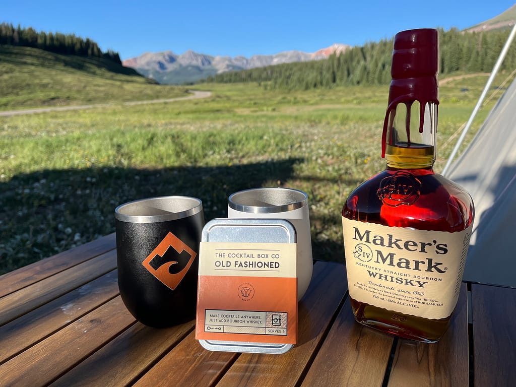 Cocktail Box Co. Old Fashioned cocktail kit with camping cocktails and a bottle of Maker's Mark at a campsite.